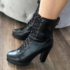 Close-up of a person wearing Aadijay black vegan leather boots by Bailaqui with rubber soles and chunky block heels on a wooden floor. The boots have silver eyelets, and a light-colored sofa is visible in the background.