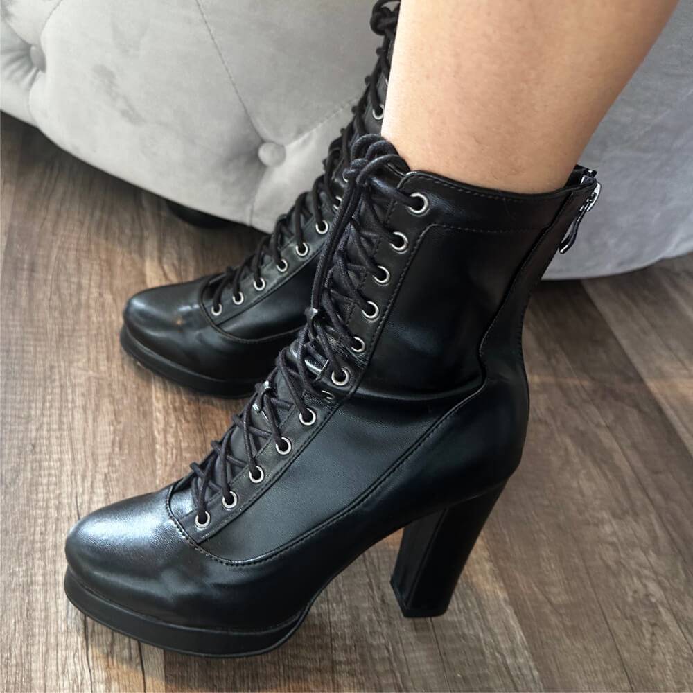 Close-up of a person wearing Aadijay black vegan leather boots by Bailaqui with rubber soles and chunky block heels on a wooden floor. The boots have silver eyelets, and a light-colored sofa is visible in the background.