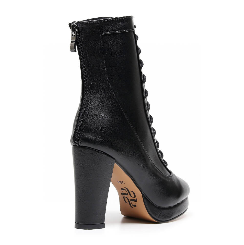 The Aadijay by Bailaqui is a high-heeled black vegan leather boot with a tan rubber sole and zipper at the back, showcasing its chunky block heel from the rear angle.